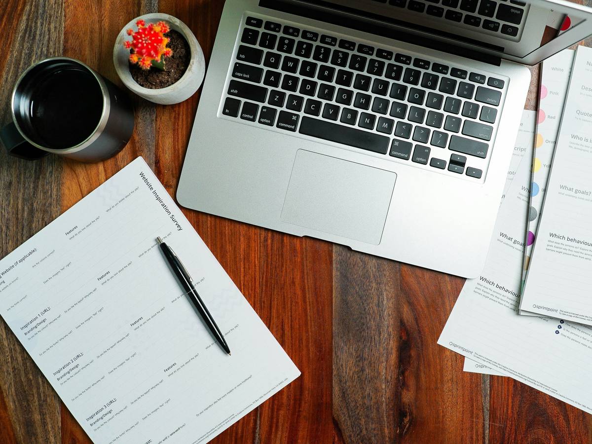 A laptop on a wooden desk among printed reports and a pen
